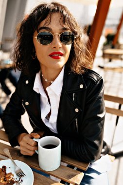 Stylish cute woman with short hairstyle with red lips wearing black jacket and white shirt is drinking coffee outdoor on summer city terrace with happy smile. High quality photo