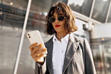 Kırmızı dudaklı, kısa saçlı ve güneş gözlüklü muhteşem güzel bayanın fotoğrafı modern şehir binasının arka planında kaydırmalı akıllı telefondur. Akıllı telefon kullanan gömlekli ve ceketli kadın portresi