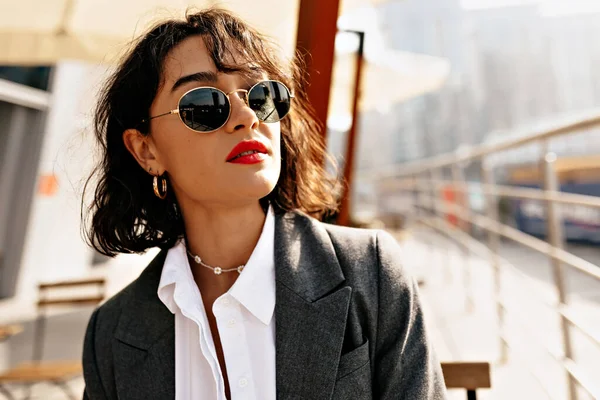 Açık tenli esmer, kısa saçlı, güneş gözlüklü, şehir arka planında poz veren kırmızı dudaklı. Kumral, dalgalı saçlı, beyaz gömlekli ve gri ceketli. Tarım kavramı.