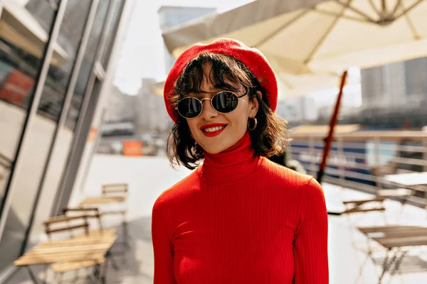 Açık havada gülümseyen güzel, genç bir kadının yakın plan portresi. Kırmızı şapkalı esmer kadın, güneş gözlüklü kırmızı bluz. Kadın bahar moda konsepti