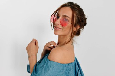 Çekici Avrupalı, bronz tenli, yuvarlak pembe gözlüklü bir kızın yakın portresi. Arkasından kameraya güzel bir gülümsemeyle poz veriyor. 