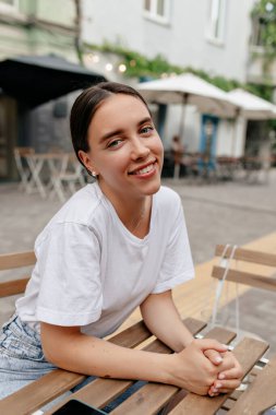 Siyah saçlı, beyaz tişörtlü sevimli gülümseyen bayan kafeteryada dinlenirken kameraya gülümseyerek poz veriyor. Yüksek kalite fotoğraf