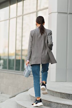 Şık ceketli ve kot pantolonlu bir bayanın arka tarafından şehirdeki cam binaya karşı merdivenlerde yürürken çekilmiş. Tarz sahibi tapılası bayan işe yürüyerek gider.. 