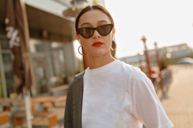 Siyah güneş gözlüklü, altın küpeli, beyaz tişörtlü ve güneşli caddede kameraya poz veren zarif bir bayan.