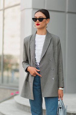 Koyu renk saçlı, kendine güveni olan, gri ceketli, güneş gözlüklü, açık havada poz veren güzel bir kadın.