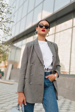 Ceketli, beyaz tişörtlü, siyah güneş gözlüklü şık bir kadın şehirdeki cam binaya karşı kameraya poz veriyor..