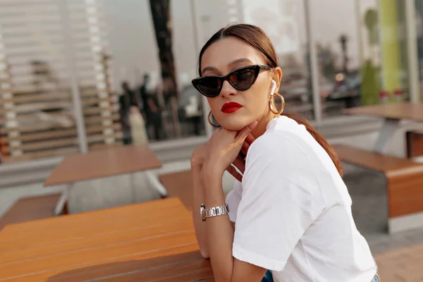 Stylish lady with dark hair in and t-shirt and dark sunglasses looks into camera in outdoor restaurant. Happy girl with bright lipstick and wrist watch in cafe.
