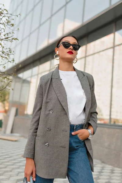 Gri ceketli, beyaz tişörtlü ve kot pantolonlu şık, genç beyaz bir kadın cam binanın arka planında el ele tutuşarak gülümsüyor. İyi ruh hali kavramı