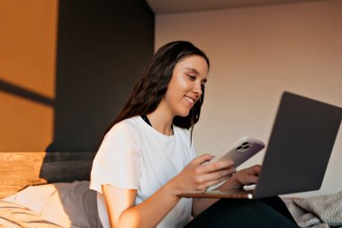 Harika gülüşü olan, dizüstü bilgisayarla çalışan ve sabah güneşinde yatakta gülümseyerek akıllı telefonu tutan muhteşem bir kız. Yüksek kalite fotoğraf