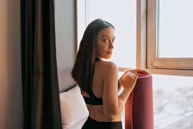 Yakışıklı, formalı, güneş ışığında pencereye karşı poz veren ve yoga minderini tutan çekici bir kadının portresini yakından çek.. 