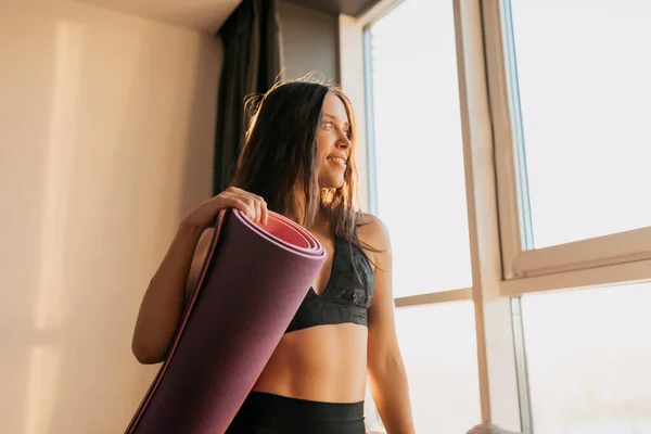 Brunette lady is standing near the window in sunshine with yoga mat in living room. Fitness healthy and sport concept. European girl preparing for morning yoga