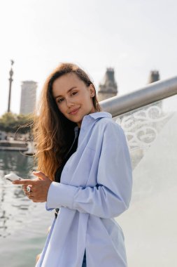 Güneşli bir günde rıhtımda akıllı telefon kullanarak mavi tişörtlü, dalgalı saçları olan çekici Avrupalı genç bir kız. Elinde akıllı telefon tutan, gülümseyen model kadın portresi. Sağlıklı yaşam tarzı. 