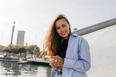 Harika gülümsemeli mutlu kız, güneşli yaz günlerinde gölün arka planında akıllı telefon kullanırken kameraya bakıyor.. 