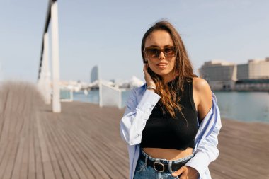 Uzun dalgalı saç stili, siyah bluzlu, güneş gözlüklü, güneş ışığında gölün arka planında poz veren hoş görünümlü bir kız. Yüksek kalite fotoğraf