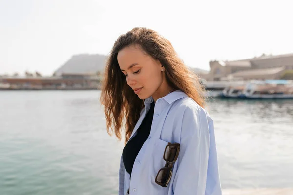Adorable pretty woman with flying wavy long hairstyle wearing blue shirt is looking down with wonderful smile while walking on pier in sunlight in warm sunny day. Resting and relaxation concept.