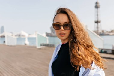 Koyu uzun dalgalı saçlı şık genç kız yaz tatilinin tadını çıkarıyor. Güneş gözlüklü ve mavi gömlekli neşeli bir öğrenci iskelede yürüyor. Rahatlama kavramı.