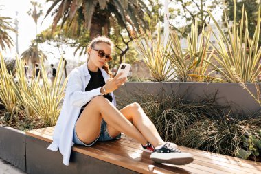 Güneş gözlüklü, mavi tişörtlü ve şortlu şık bir kızın tam boy fotoğrafı sokak bankında oturuyor ve güneş ışığında akıllı telefon kullanıyor.