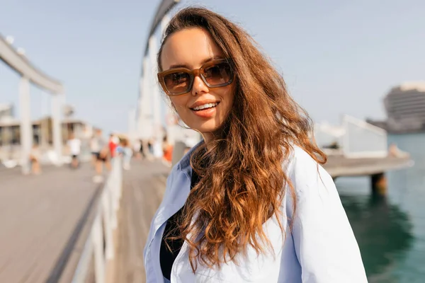 Açık havada, uzun dalgalı saç stiliyle gülümseyen güzel bir kızın yakın portresi. Kameraya gülümsüyor ve yaz tatilinin tadını çıkarıyor.. 