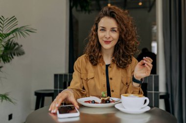 Avrupalı güzel, bukleli, kahverengi tişörtlü, modern modaya uygun bir kafede kahvaltı yapan kızın kapalı bir portresi.. 
