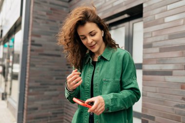 Dışarıda, yeşil tişörtlü güzel, kıvırcık bir kızın akıllı telefonu kaydırması ve dışarıda yürürken gülümsemesi. Tasasız genç Avrupalı kız dışarıda modern akıllı telefon kullanıyor.. 