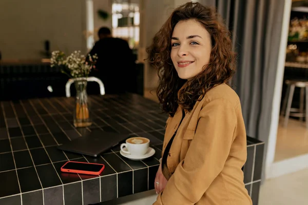 Happy excited girl with wavy hair wearing brown shirt sitting in cage with coffee and smartphone and smiling at camera. Weekend, morning, breakfast with friends