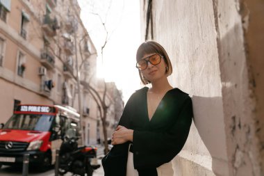 Saf, beyaz, sıradan gömlekli bir kadın aramasını bekliyor. Şehir arka planında akıllı telefon tutan rahat bir kızın açık hava fotoğrafı..
