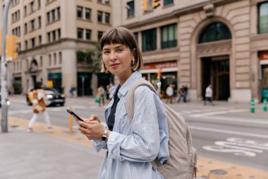 Kısa düz saçlı, mavi tişörtlü, akıllı telefon tutan ve kameraya şehrin arka planında gülümseyerek poz veren çekici bir kız.. 