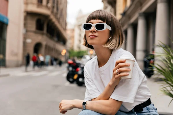 Attractive pretty lady with short hair in sunglasses wearing casual clothes spend time outdoor with coffee in the summer morning. Graceful lady holding cup of tea posing during resting outdoor.