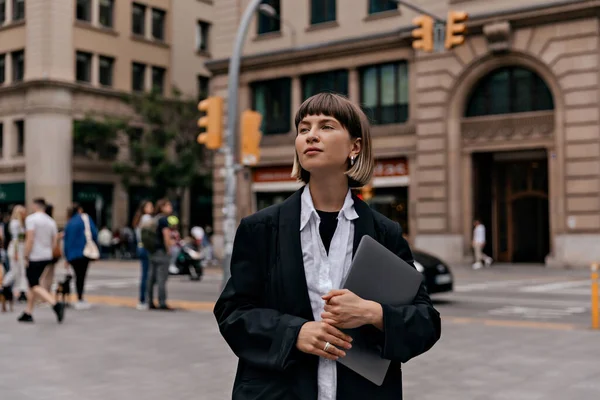 Business woman in black jacket and white shirt with straight short hair holding laptop walking in business district. Successful positive girl going after work outside.