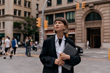 Siyah ceketli, beyaz gömlekli, kısa saçlı, dizüstü bilgisayarı olan bir iş kadını iş bölgesinde yürüyor. Başarılı pozitif kız işten sonra dışarı çıkıyor..