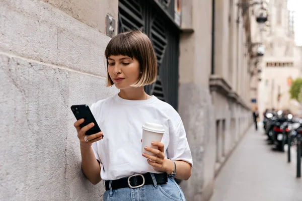 Incredible charming woman with short hair wearing white t-shirt scrolling smartphone in the city. Young charming lady with coffee and telephone in the city.