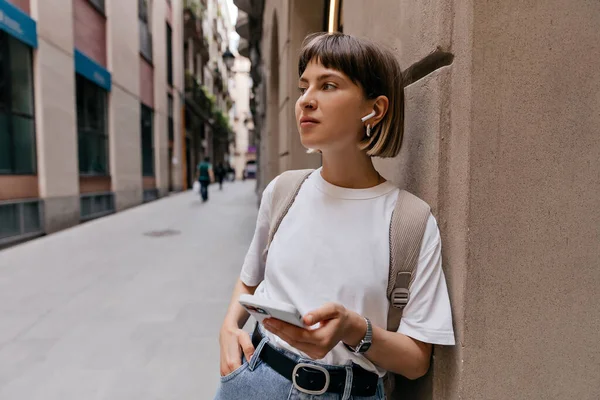 Cheerful woman with smartphone in wireless headphones smiling in city. Lady with short hair in white t-shirt, jeans jeans posing outside on old European street