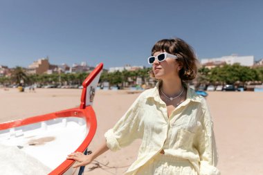 Dalgın kısa saçlı kadın güneş ışığında teknenin yanındaki kumlu sahilde takılıyor. Dışarıda poz veren sevimli bir kız. Kahverengi saçlı güneş gözlüklü kadın eğleniyor ve ilk sıcak günlerinin ve yaz tatilinin tadını çıkarıyor..