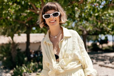 Kumsaldayken ağzı açık gülen beyaz, kısa saçlı, mutlu genç kızın yakın çekimi. Sarı gömlekli, şeffaf gözlüklü güzellik. Boş zaman, rahatlama, yaşam tarzı konsepti.