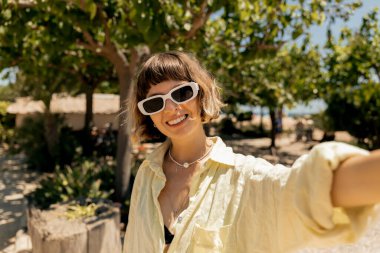 Dalgalı kısa saçlı, güneş gözlüğü takan harika bir stil sahibi kız plajdaki yeşil bitkilerin arka planında güneşli yaz gününde selfie çekiyor. 