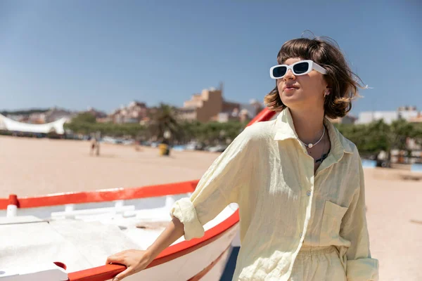夏のスタイリッシュな衣装とサングラスで愛らしい素敵な魅力的な女の子は ボートの近くのビーチで晴れた暖かい日にポーズをとっています 休暇を楽しむ概念 — ストック写真
