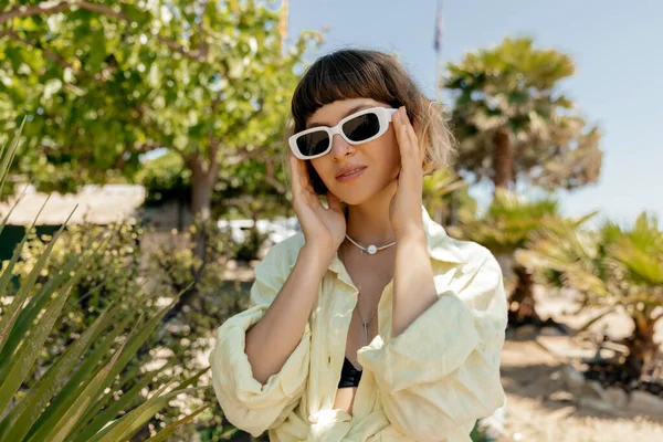 紧贴时髦女孩的肖像 她的头发短短的 戴着太阳镜 穿着夏黄西服 触摸着她的眼镜 在夏日的背景下在棕榈树上摆姿势 — 图库照片