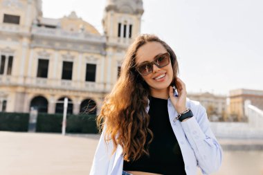 Happy young european girl laughs looking at camera, standing on pavement in middle of square on street. Brunette wears black T-shirt, jeans and blue shirt. Mood, lifestyle, concept.