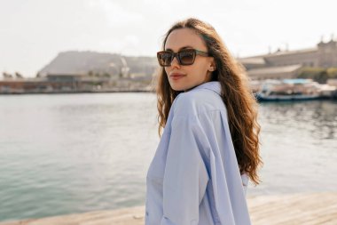 Güneş gözlüklü ve mavi gömlekli zarif çekici kadın gölün arka planında güneşte kameraya poz veriyor. Haftasonu denizde görülen şık kız. 