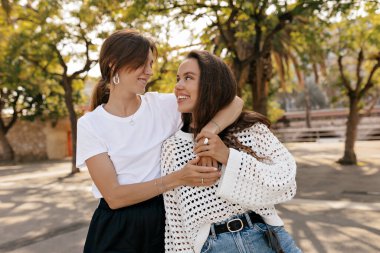 Moda kıyafetleri içinde gülen ve dışarıda yürüyen iki esmer kız. Kot şortlu ve çiçekli bluzlu çekici kadınlar gülümseyip konuşuyor..