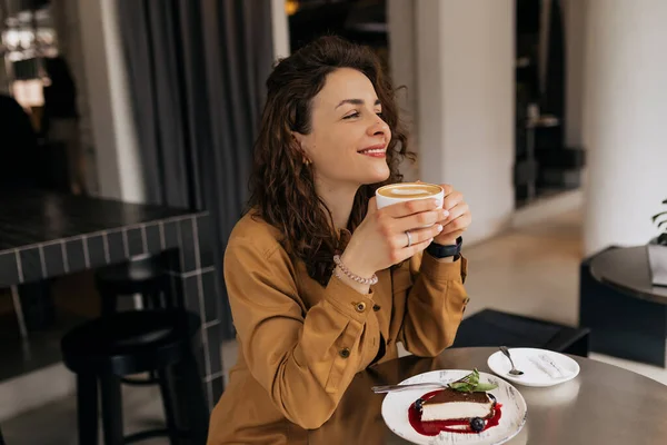 Happy charming lady with curls wearing brown shirt enjoying morning coffee and dessert in cafe. Caucasian stylish lady is having breakfast in cafe.