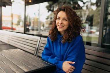 Kıvırcık mavi tişörtlü hoş bir bayan yaz için ahşap terasta oturuyor ve arkadaşlarını bekliyor. Çekici gülümseyen kızın açık hava portresi 