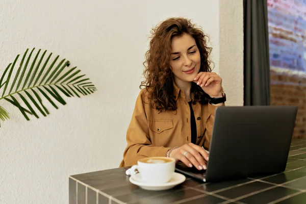 Stylish pretty woman with curly hair wearing brown shirt sitting in cafe with coffee and working on laptop. European girl in stylish outfit in modern city cafe.