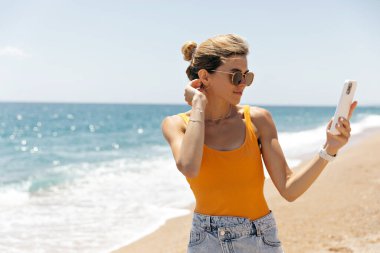 Fashionable girl with collected hair, tanned skin and great smile wearing orange t-shirt and denim shorts is making selfie on the beach in sunny hot day. 