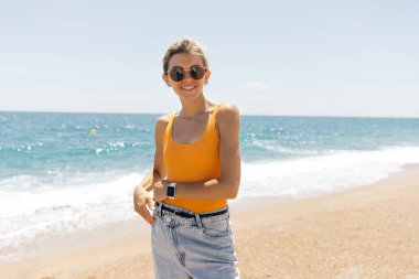 Sahilde arka planda deniz pozu veren çarpıcı bir kadın. Güneş gözlüklü muhteşem genç kadın doğanın arka planında fotoğraf çekilirken gülümsüyor..