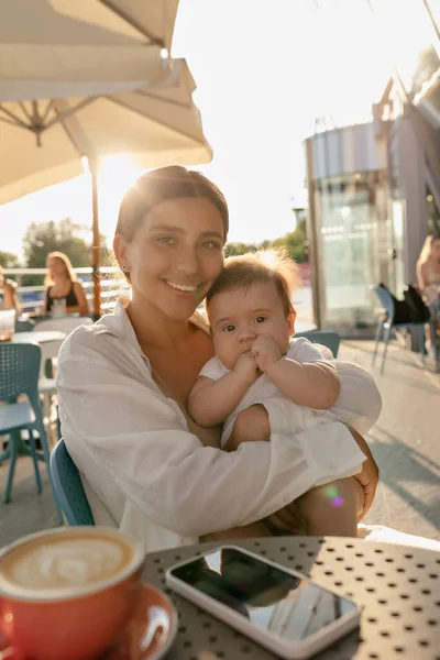 Outdoor portrait of happy young mom with baby boy posing with smile on summer terrace in sunlight. Charming woman relaxing in the city with her son on sunrise