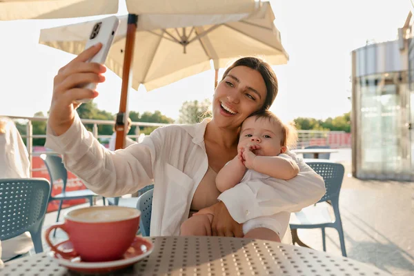 Happy excited lovely woman with tanned skin and wonderful smile wearing white shirt resting on terrace in sunlight with baby and making selfie. Happy young mom spend weekend with her son.