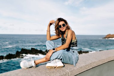 Avrupa tarzı tişörtlü ve kot pantolonlu bir kadının okyanus dalgalarının arka planında güneş ışığıyla taş sahilde oturduğu uzun bir açık hava fotoğrafı. Kumsalda dinlenen mutlu kadın 