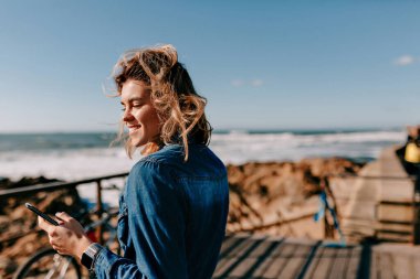 Smiling happy european woman wearing denim shirt standing on ocean shore with on ocean waves background and looking straight while listening music 