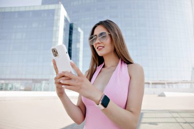 Güneş gözlüklü, pembe yazlık elbise giyen, akıllı telefon kullanan ve güneşli bir günde şehirde dolaşan inanılmaz şık bir kadın..